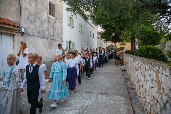 Djeca u nošnjama u povorci niz ulicu između kamenih zidova.