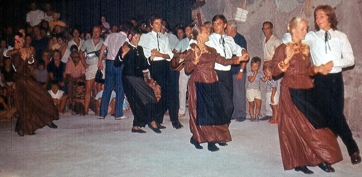 Stara fotografija: plesačice u dugim smeđim i tamnim nošnjama plešu s plesačima pred mještanima.